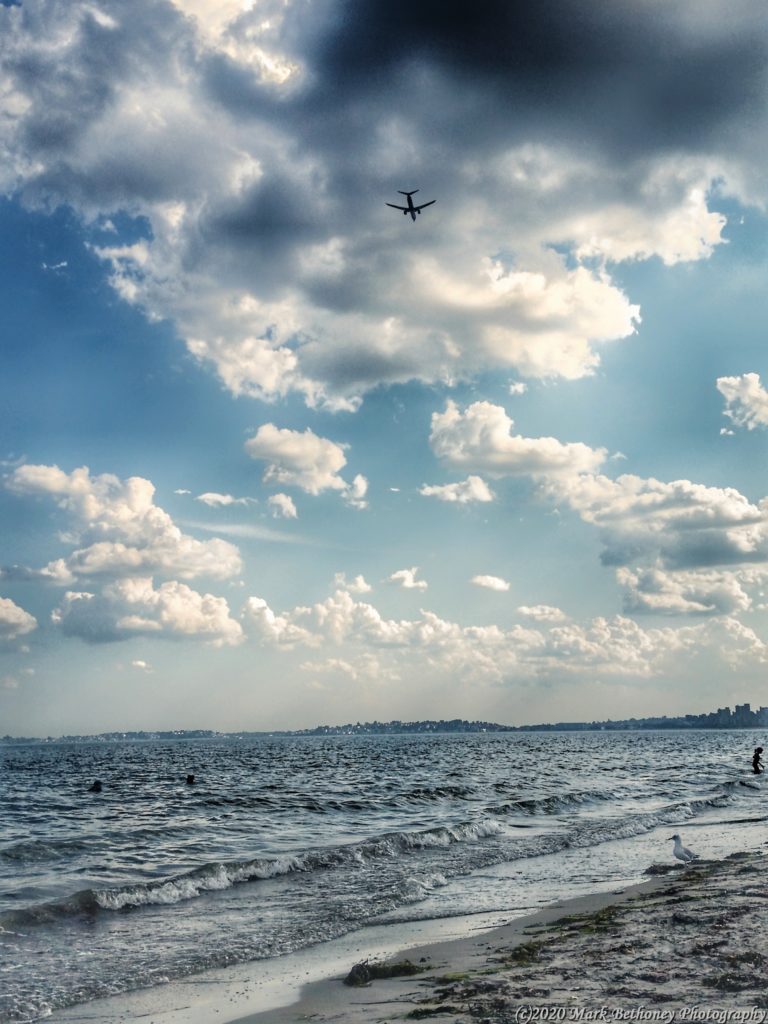 A shot of Revere Beach with the sand and water in the foreground and an airplane flying overhead into the clouds.